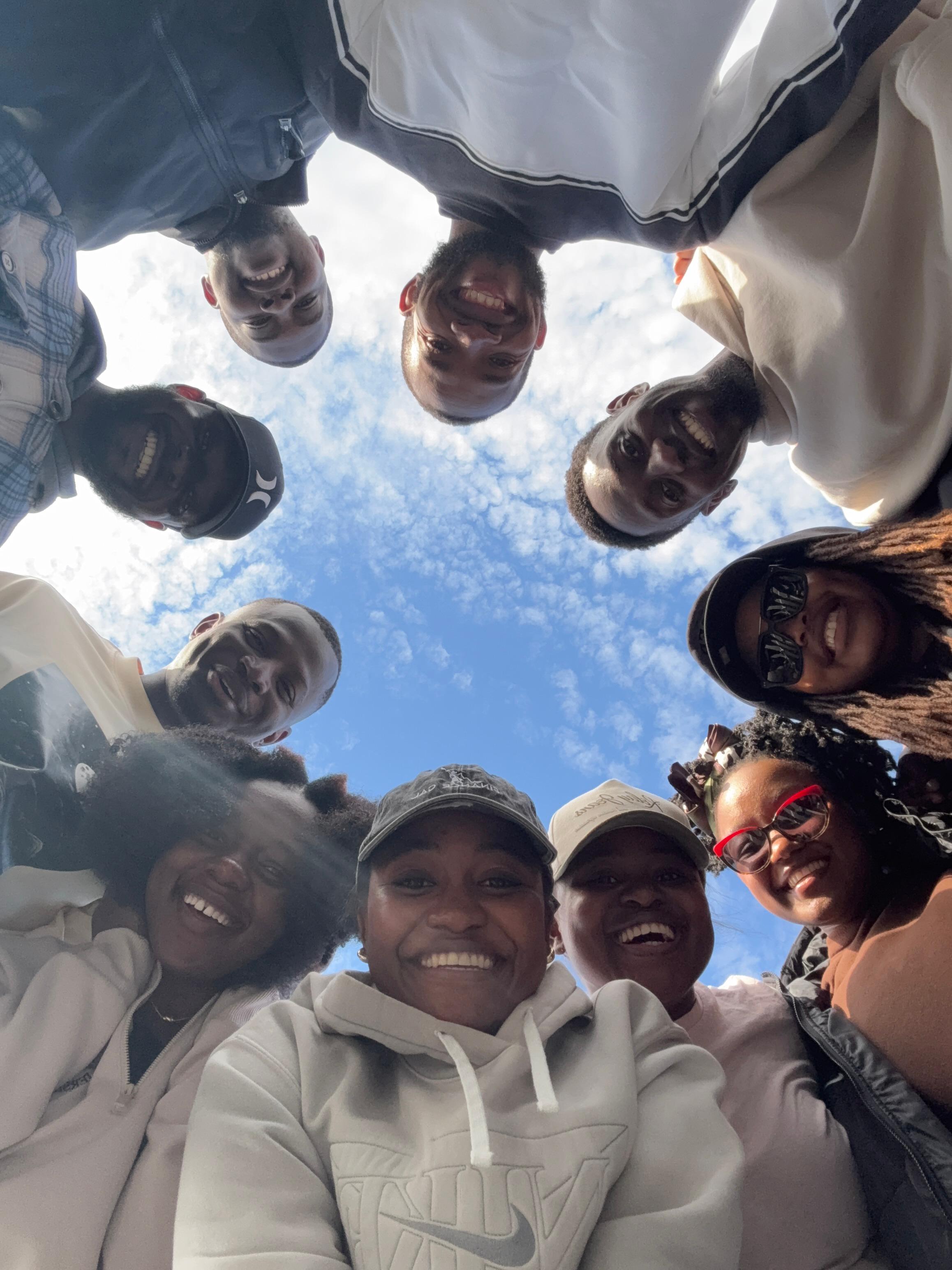 MAGIS members standing in a circle looking down at the camera under a blue sky